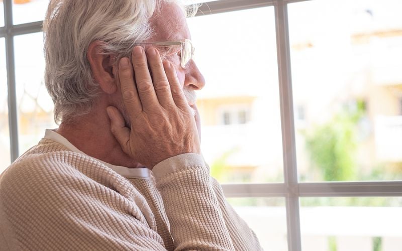 elderly man staring out of the window looking worried.